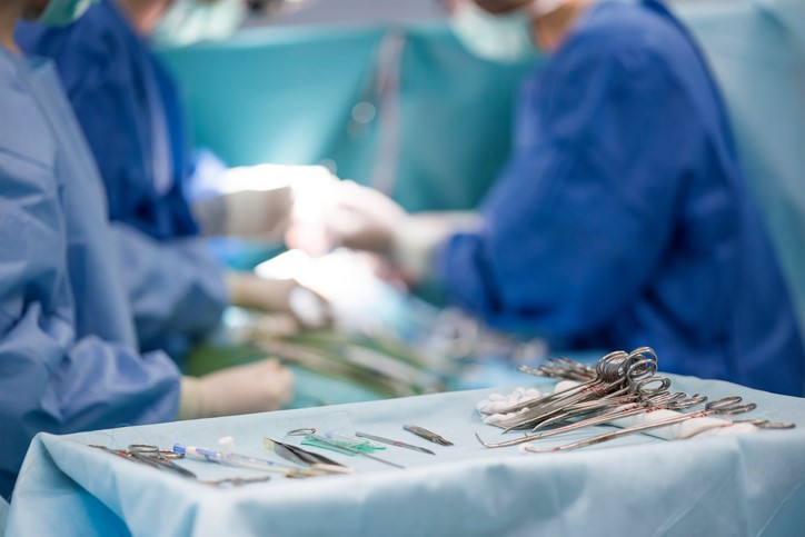 Picture of a faded background of a Surgeon Team handing Medical Instruments to the Surgeon. There is a focus image shot of Medical Instruments sitting on top of a table.