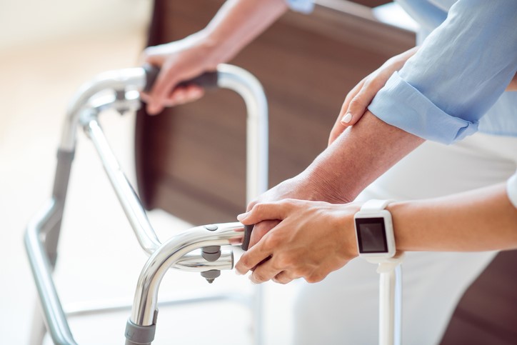 Picture of a patient's hands holding on to a walker in front of a swing bed. There is someone else's hands on patients while they assist them with the walker.