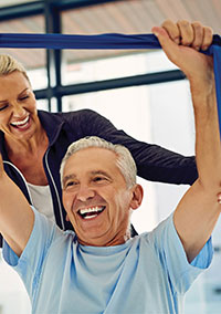 Picture of a smiling older man lifting his arms up while using an exercise stretch band. There is a woman smiling and standing behind him as she helps assist him with his strength.